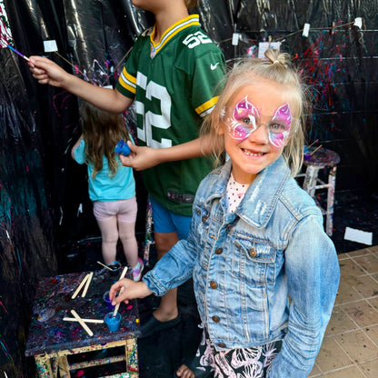 Children with face paint standing in front of a black backdrop with colorful paint splatters.
