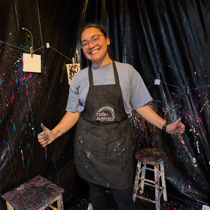 Instructor wearing an apron in a studio with art supplies and black curtains.