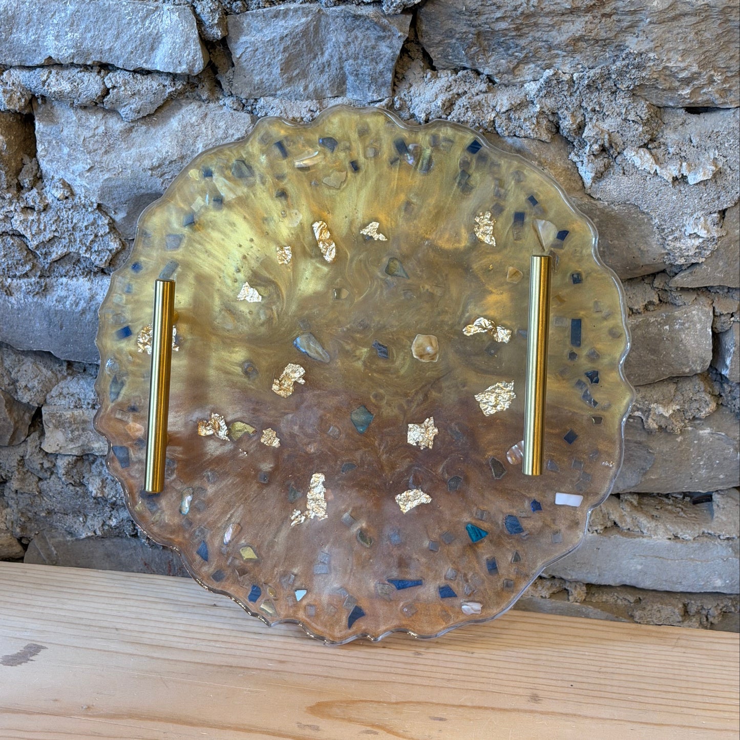 A round, scalloped-edge tray made of translucent epoxy with metallic gold flakes, shown against a rustic stone and wood background.