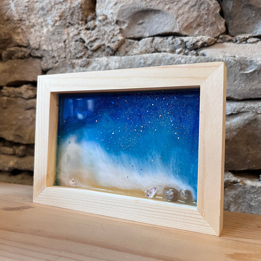 A wood frame containing a blue and white epoxy beach design with small clear shells, displayed on a wooden surface against a rustic stone wall.