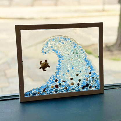 A framed glass art piece depicting a blue ocean wave with a small sea turtle, displayed on a dark windowsill with a blurred outdoor background.