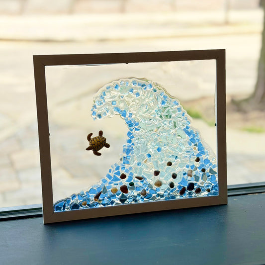 A framed glass art piece depicting a blue ocean wave with a small sea turtle, displayed on a dark windowsill with a blurred outdoor background.