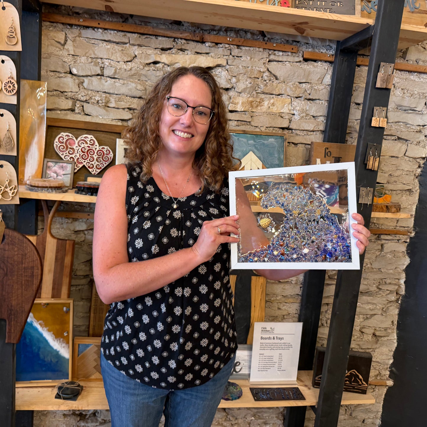 A woman in a studio holding up framed glass art. The artwork features a blue wave design with a turtle.