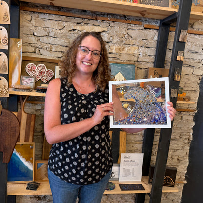 A woman in a studio holding up framed glass art. The artwork features a blue wave design with a turtle.