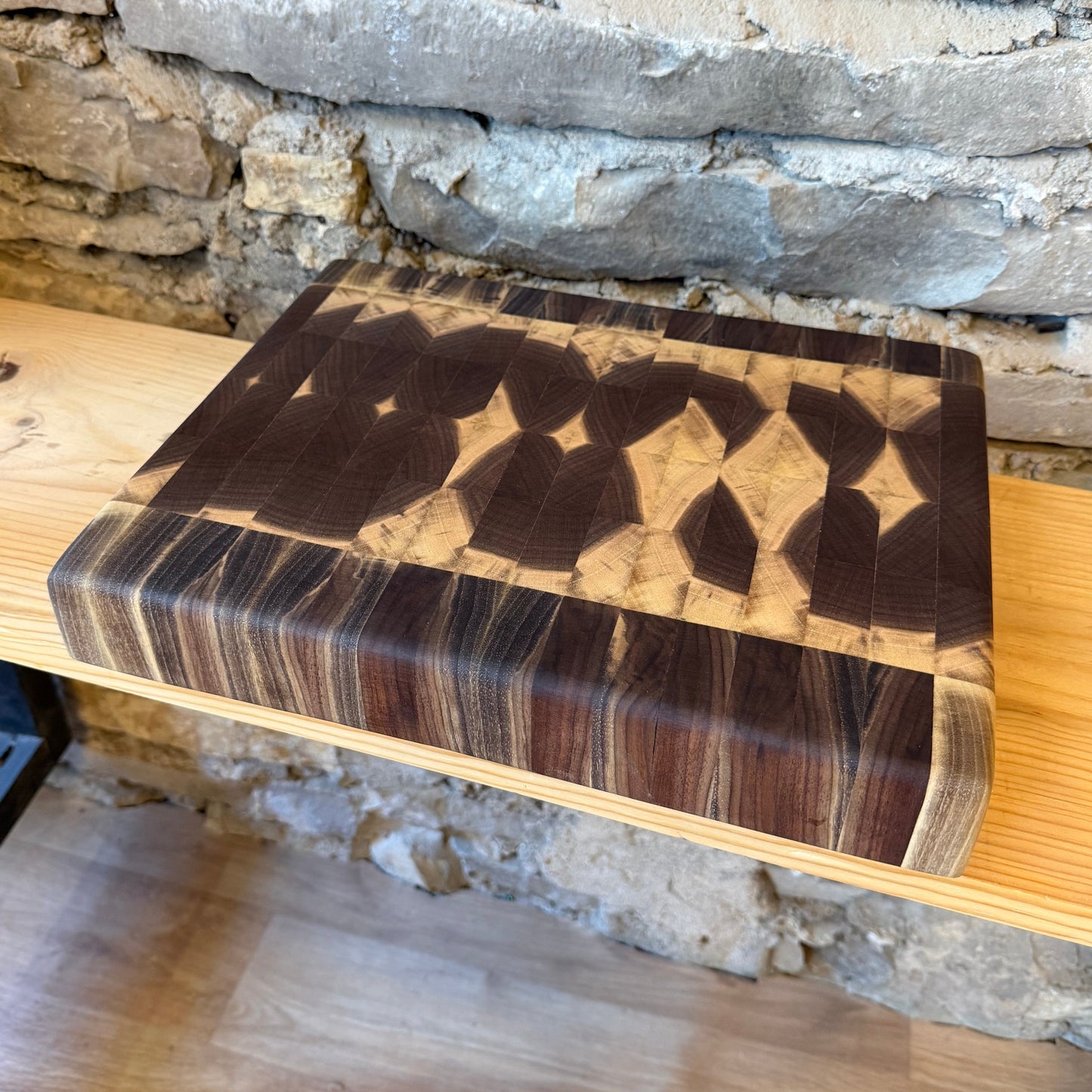 A rectangular wooden cutting board with a geometric inlay pattern, resting on a light wood shelf in front of a rustic stone wall.