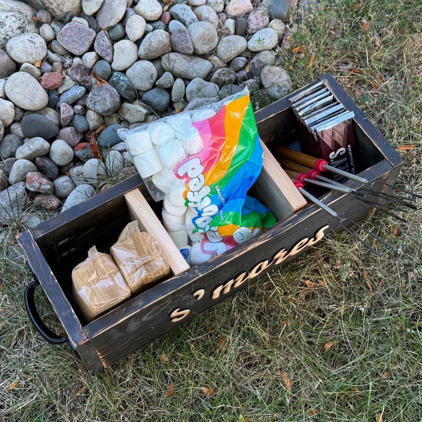 A wooden s'mores tray filled with marshmallows, chocolate, graham crackers, and stick, placed on grass next to a fire ring of rocks.