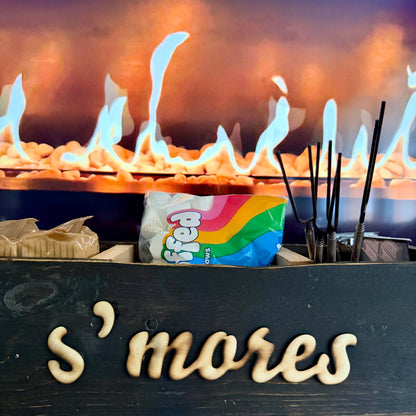A wooden s'mores tray in front of a fire, holding a bag of marshmallows, chocolate, graham crackers, and sticks. 