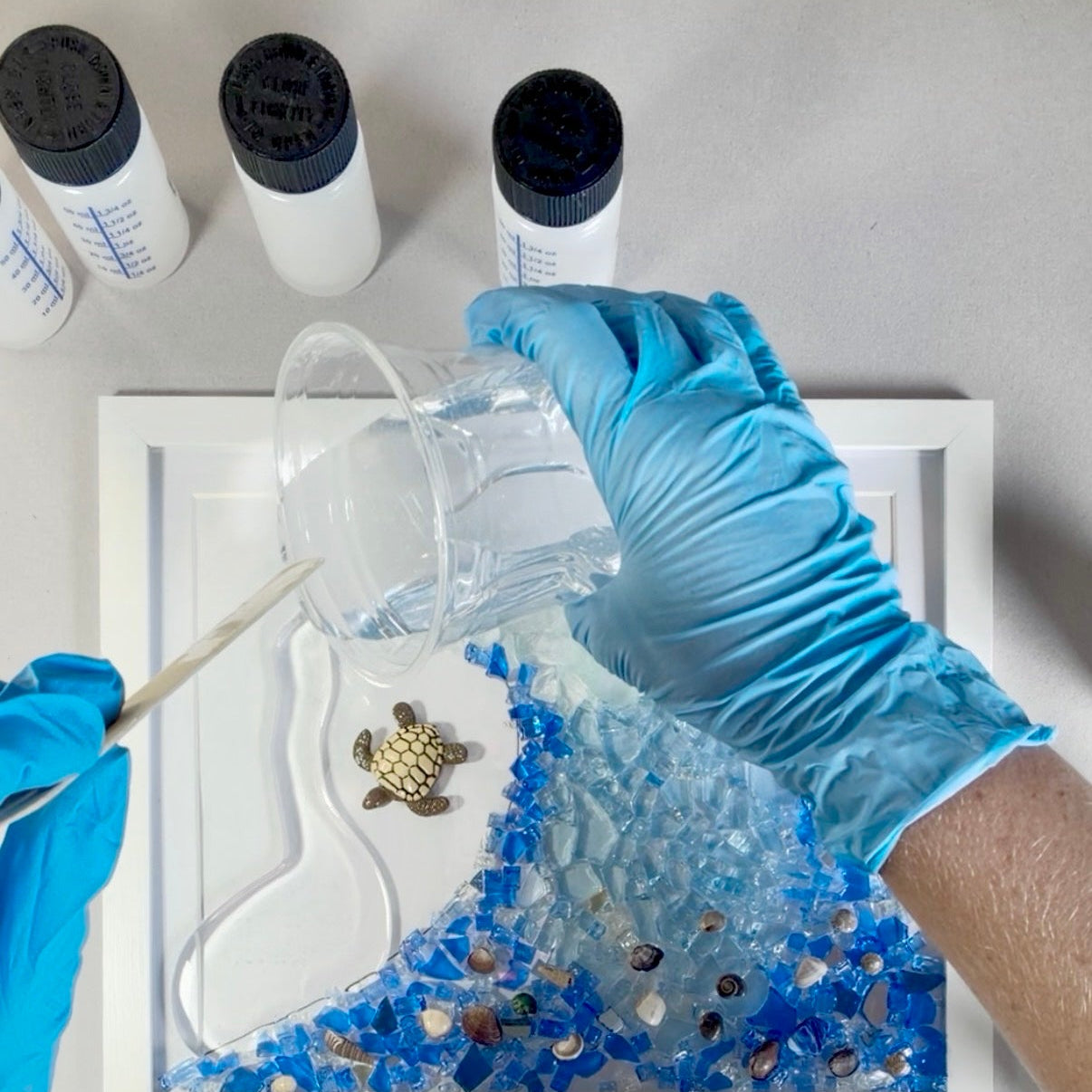 A top-down view of a person wearing blue gloves pouring clear resin over a white frame filled with blue glass and a small sea turtle charm, with bottles and tweezers on a white surface.