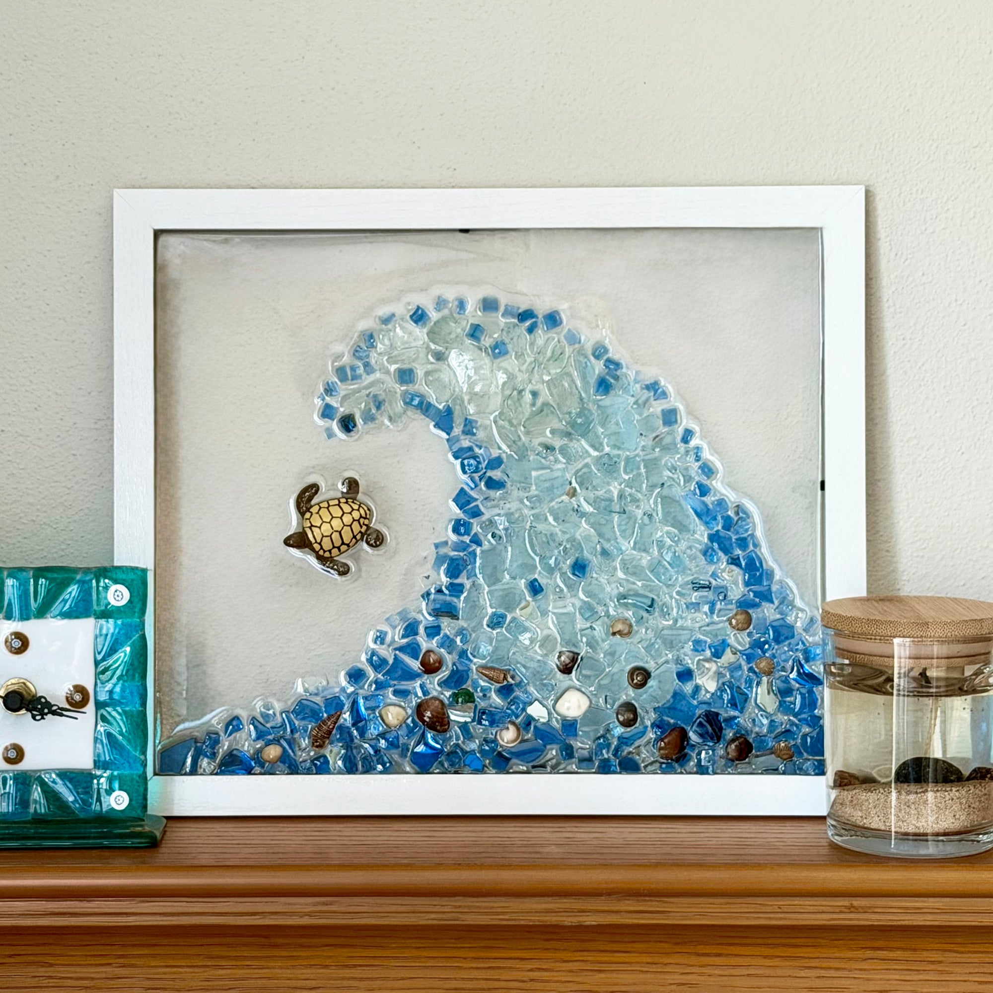 A framed mosaic art piece featuring a blue glass wave and a small sea turtle, displayed on a wooden shelf next to other home decor items.