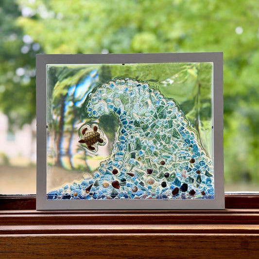 A square framed glass mosaic art piece depicting a blue and white ocean wave with a small turtle, displayed on a wooden windowsill with a blurred green garden background.