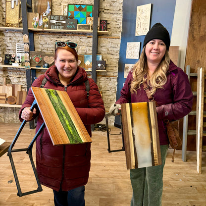 Two women holding wood and epoxy tables with artistic designs in a studio setting.