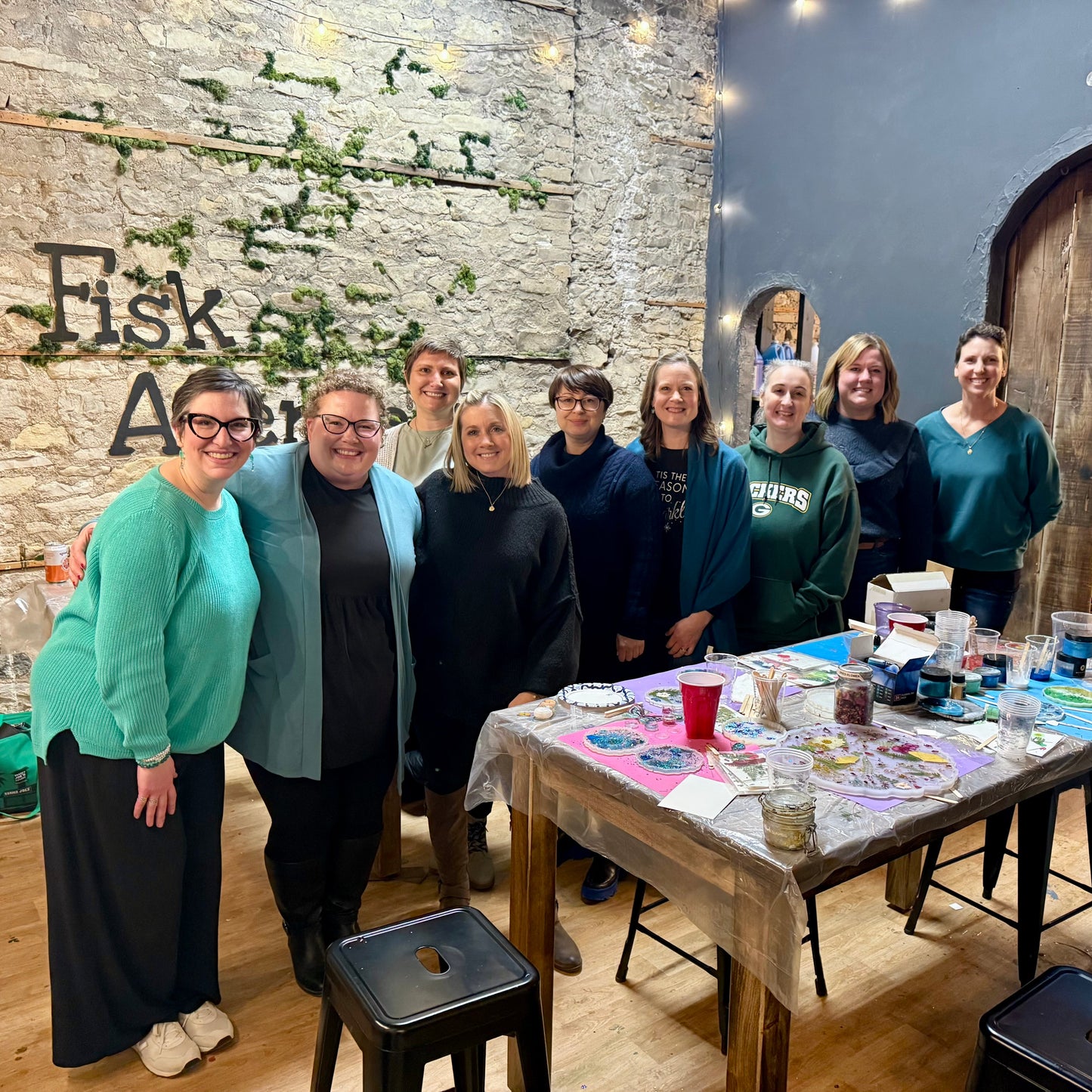 Group of people posing in front of a stone wall with 'Fisk Art' sign, table with art supplies in the foreground.