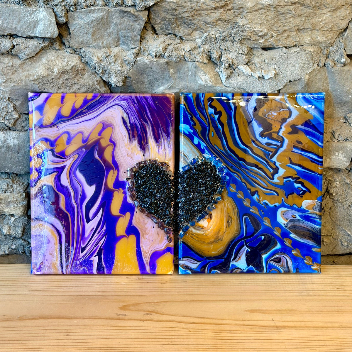 Two square abstract wall art canvases with fluid marbled patterns, displayed side-by-side on a light wood surface against a rustic stone wall. The left canvas features purple, blue, and gold swirls with a black textured heart in the center.