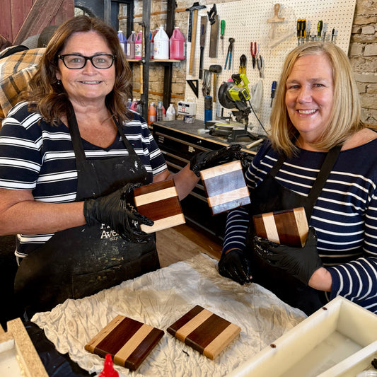 Two women in a workshop holding wooden coasters, surrounded by tools and materials.
