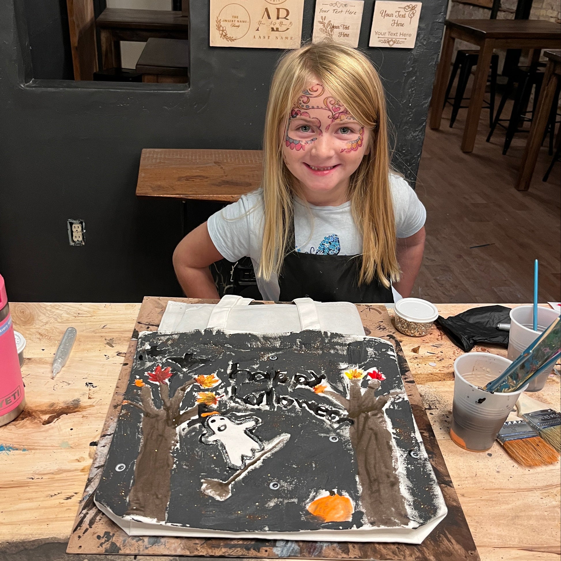 Person in a workshop with art supplies and a painted trick-or-treat tote bad on a table.