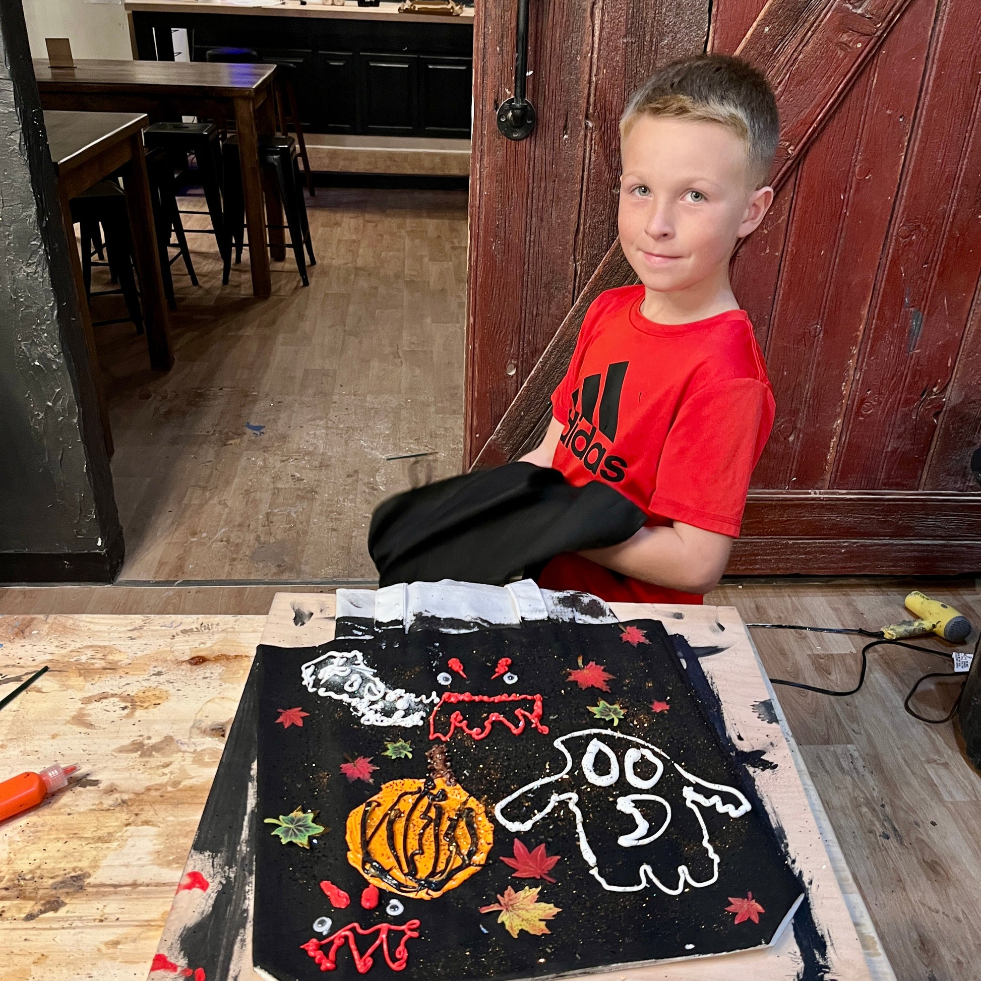 Child in a red shirt sitting behind a wooden table with a painted trick-or-treat tote bag, in a studio setting.