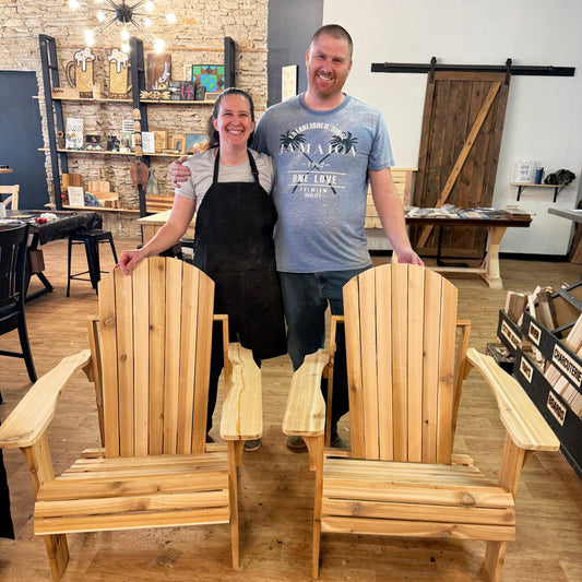 Two people standing behind two wooden Adirondack chairs in a workshop setting.