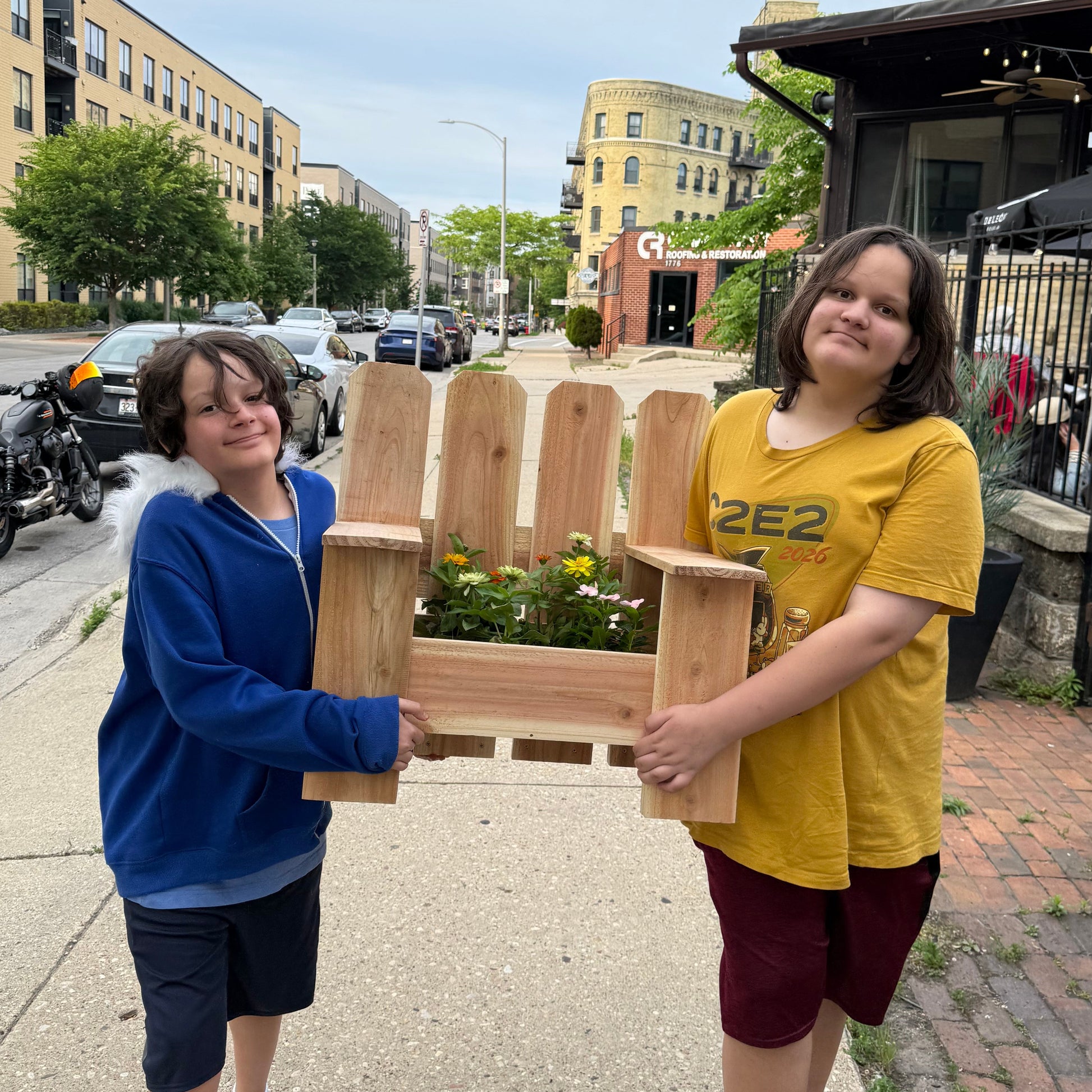 Two people standing on a sidewalk holding a planter chair with flowers and a cityscape in the background