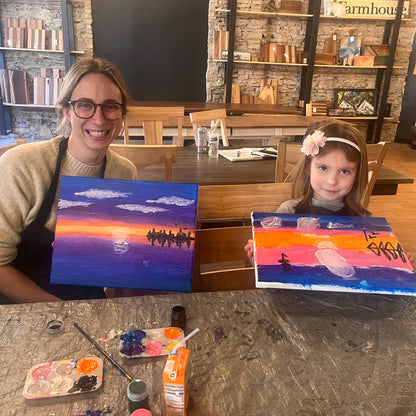 Woman and child holding paintings of sunsets with trees in a room with shelves and furniture.