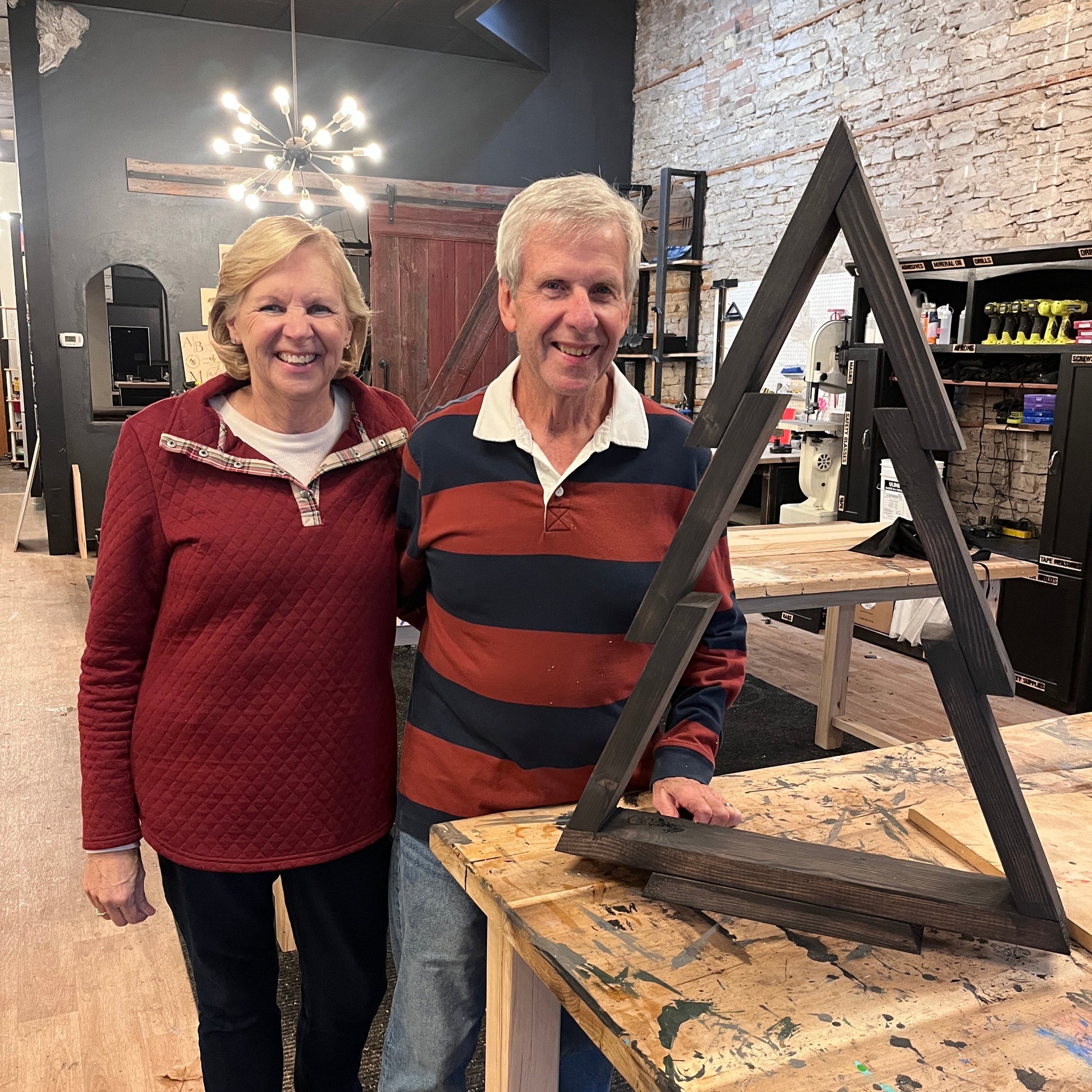 An older couple standing in a workshop setting, smiling and posing with a decorative pine tree craft that they have created.