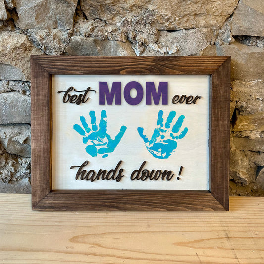 Framed artwork with handprints and text 'best MOM ever hands down!' against a stone wall background