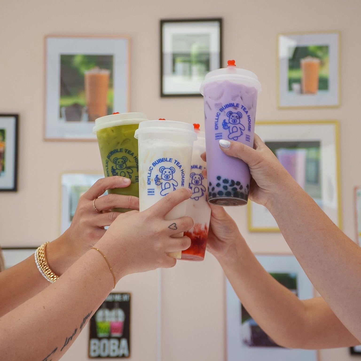 Four hands holding bubble tea cups in front of a wall with framed pictures of drinks.