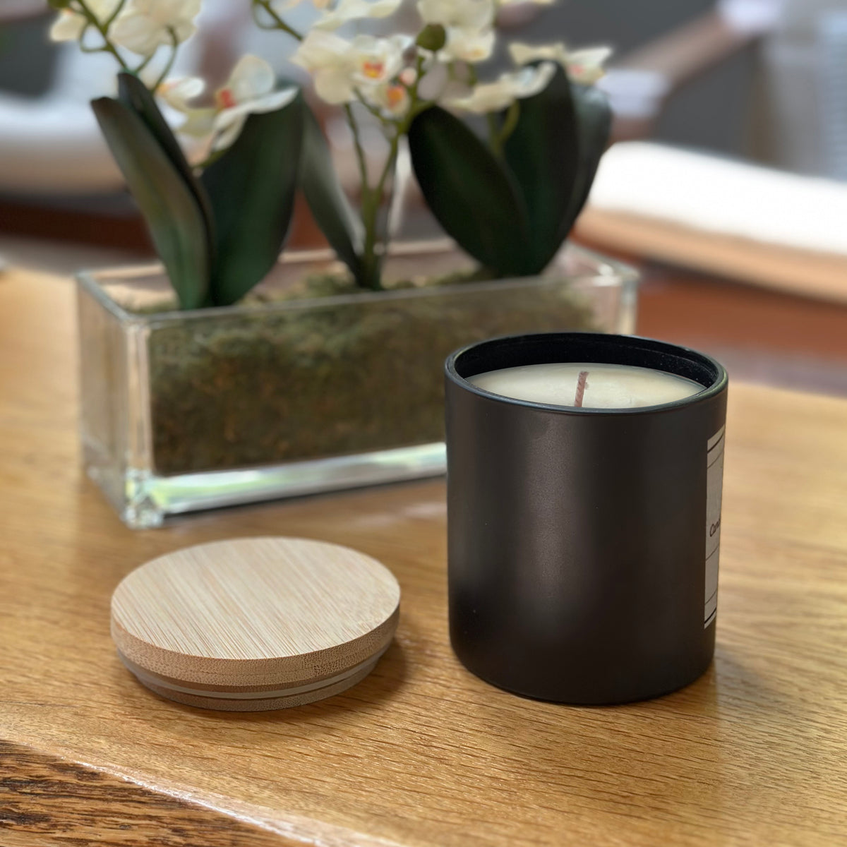 Black candle with wooden lid on a wooden surface, with a vase of flowers in the background.