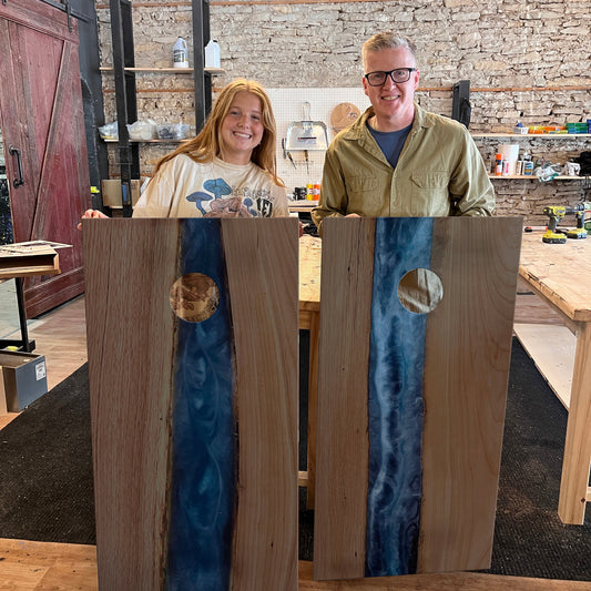 Two people standing behind wood and epoxy cornhole boards with blue inlays in a studio setting.