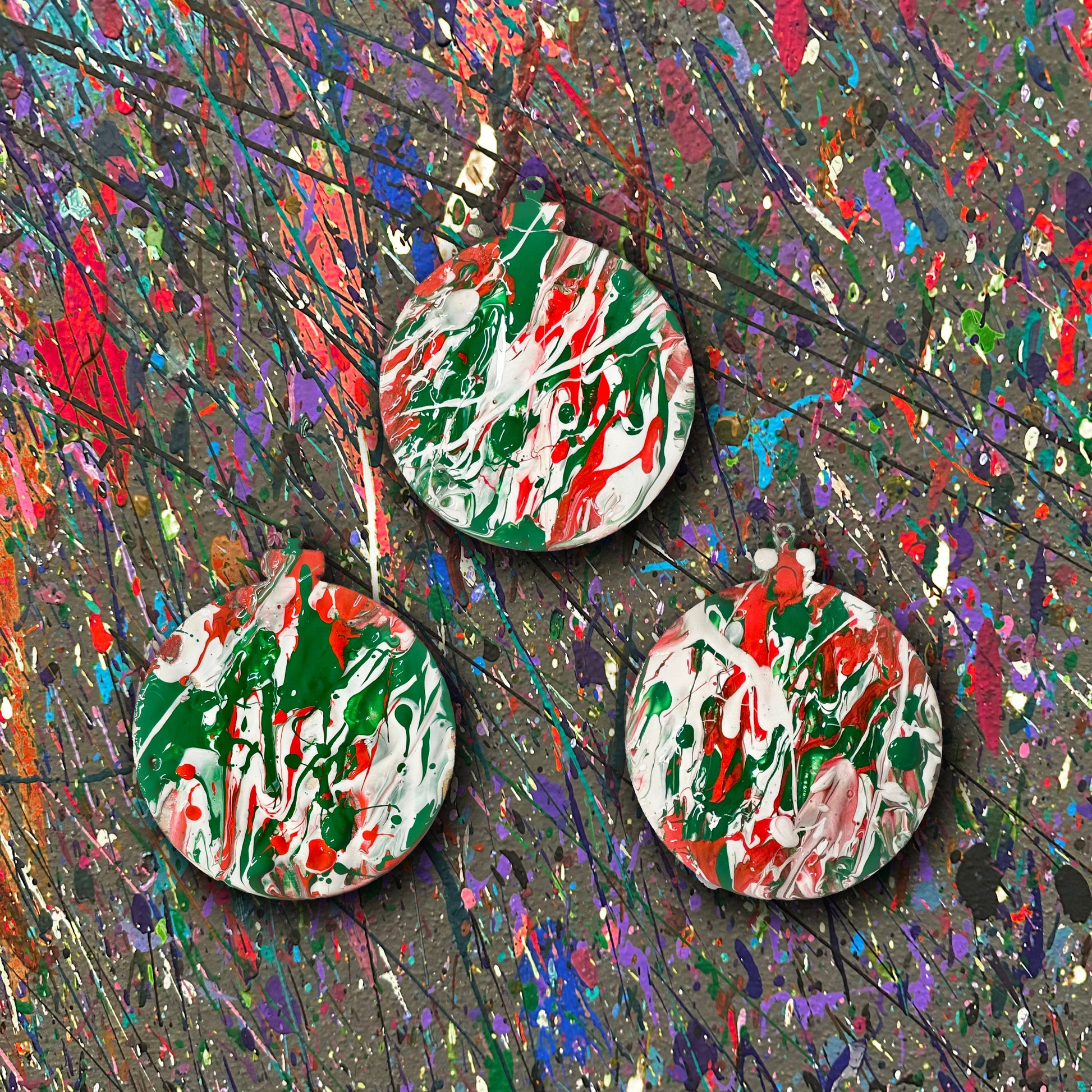 Set of three colorful splatter painted ornaments on a textured surface with splashes of red, green, and white.