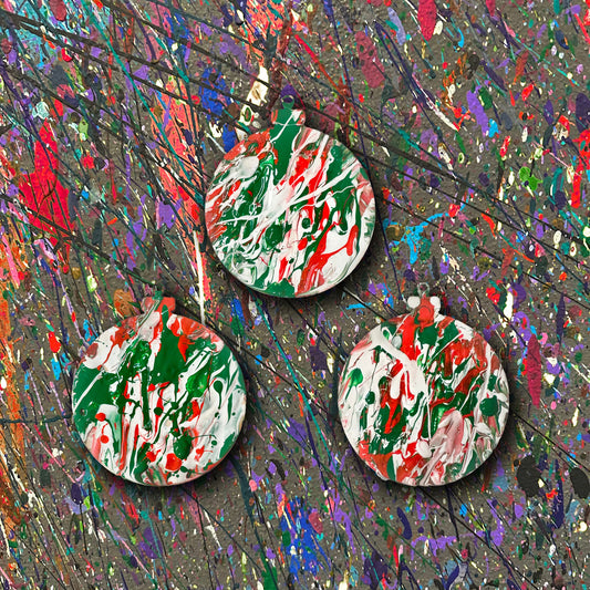 Set of three colorful splatter painted ornaments on a textured surface with splashes of red, green, and white.
