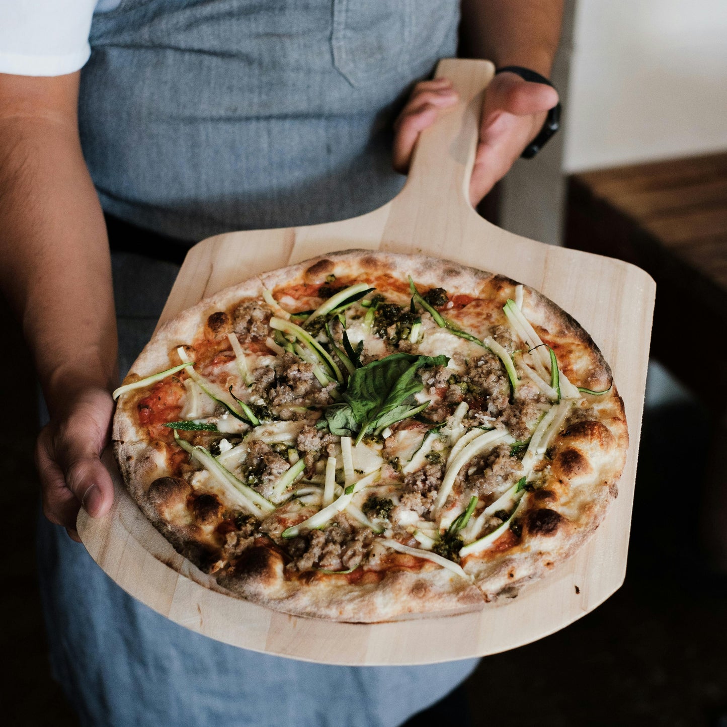 Person holding a pizza on a wooden pizza peel board with a blurred background