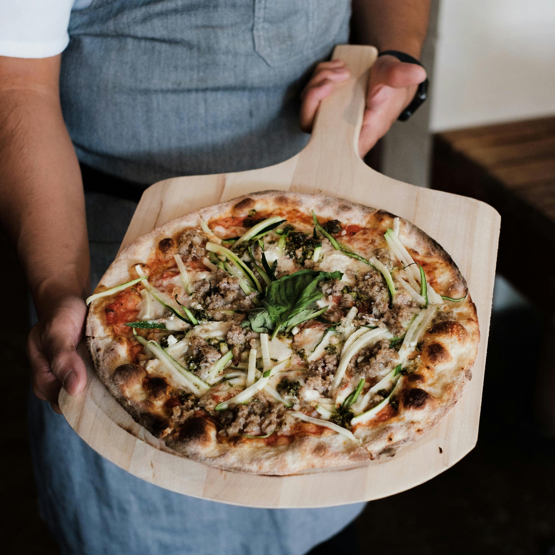 Person holding a pizza on a wooden pizza peel board with a blurred background