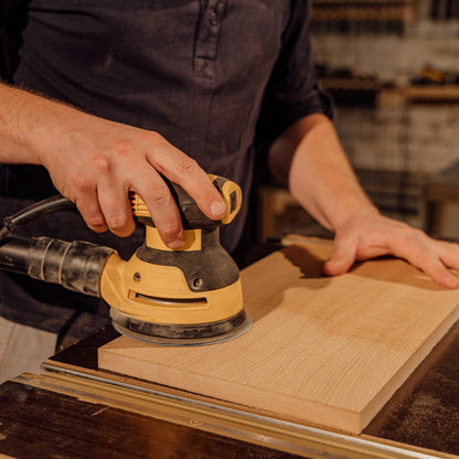 A person is using a power sander tool to work on a piece of wooden furniture in a workshop setting.