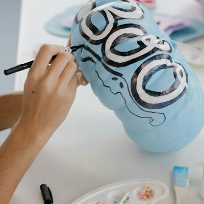 Person painting a blue pumpkin with the word 'BOO' and black ghosts designs on a white surface.