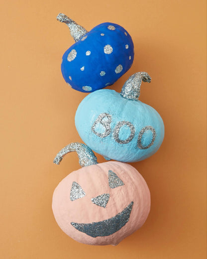 Three painted pumpkins with a blue polka-dotted pattern, the word 'B00' on a light blue background, and an orange jack 'o lantern face, placed on an orange surface.