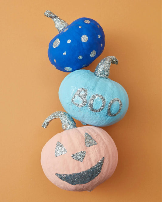 Three painted pumpkins with a blue polka-dotted pattern, the word 'B00' on a light blue background, and an orange jack 'o lantern face, placed on an orange surface.