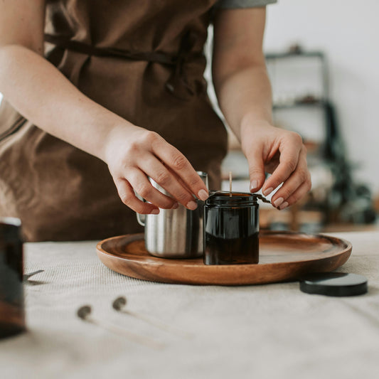 Person in a brown apron preparing a handmade candle on a wooden tray in a studio setting.