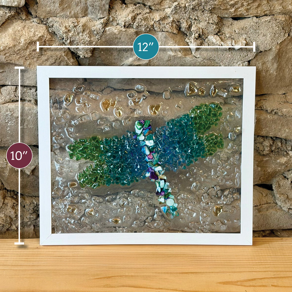 Framed dragonfly wall art made from colorful glass pieces, shown leaning against a stone wall on a wooden surface. White dimension markers indicate 12 inches wide and 10 inches tall.