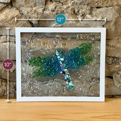 Framed dragonfly wall art made from colorful glass pieces, shown leaning against a stone wall on a wooden surface. White dimension markers indicate 12 inches wide and 10 inches tall.