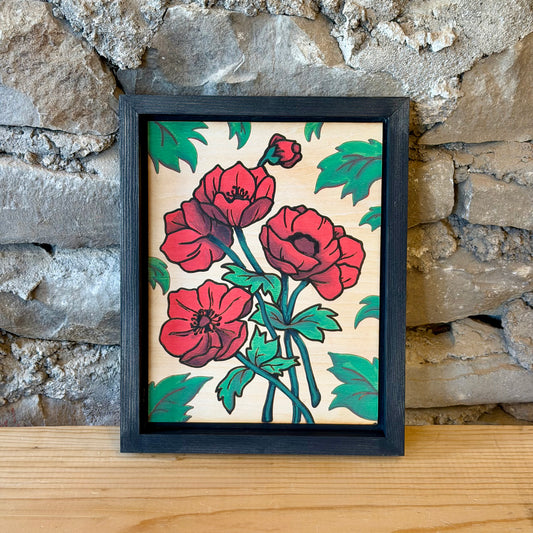 A framed art print featuring red poppy flowers and green leaves on a natural wood background, set in a black frame and leaning against a rustic stone wall on a wooden surface.
