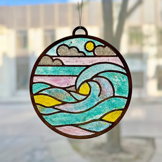 A circular stained-glass-style hanging ornament depicting ocean waves, clouds, a sun, and mountains, suspended in front of a softly blurred outdoor window background.