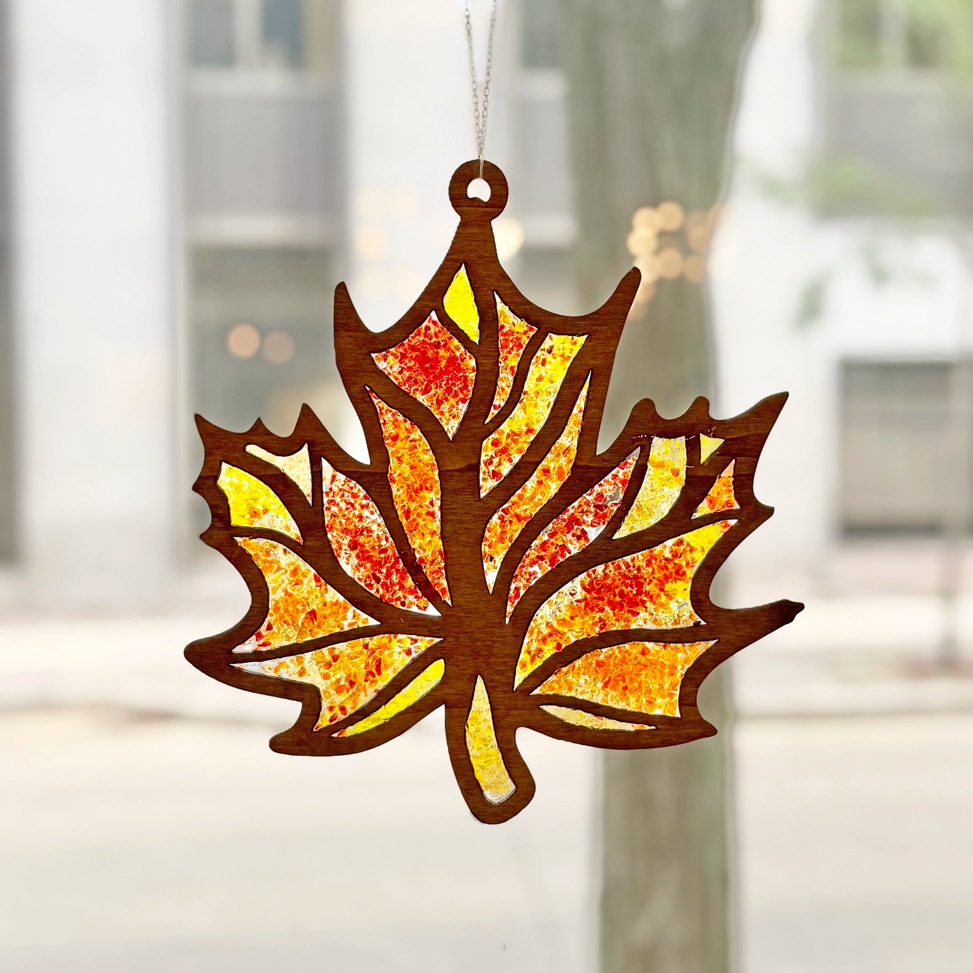 A laser-cut autumn maple leaf suncatcher with a warm brown wood outline and a glass yellow, orange, and red patterned center, hanging in front of a softly blurred window background.