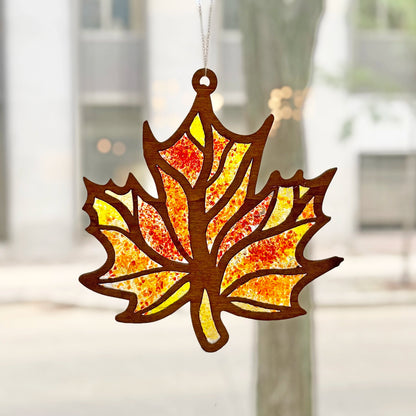 A laser-cut autumn maple leaf suncatcher with a warm brown wood outline and a glass yellow, orange, and red patterned center, hanging in front of a softly blurred window background.