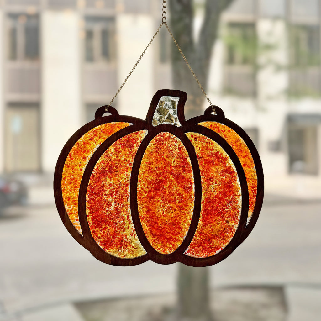 A pumpkin-shaped stained glass suncatcher with a textured orange and yellow design, hanging from a clear suction cup on a window in front of a blurred outdoor city background.